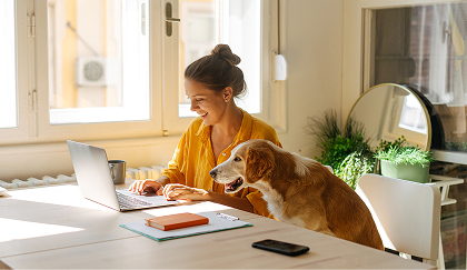 A woman and her dog scroll on a laptop while seated at a kitchen table