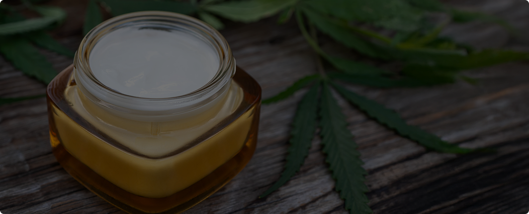 Open jar of topical CBD product surrounded by cannabis leaves on a wooden surface